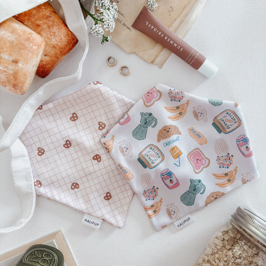 Two fabric face masks on a white surface with coffee and breakfast foods